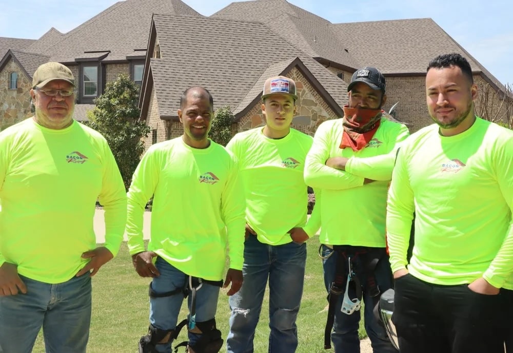 Five roofers in neon yellow Bacon Roofing shirts posing on the grass outside a brick house